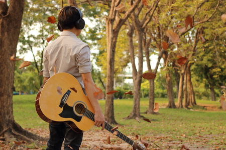 Rear view of portrait of handsome young man holding acoustic guitar with headphones against among falling leaves in the park outdoorsの写真素材