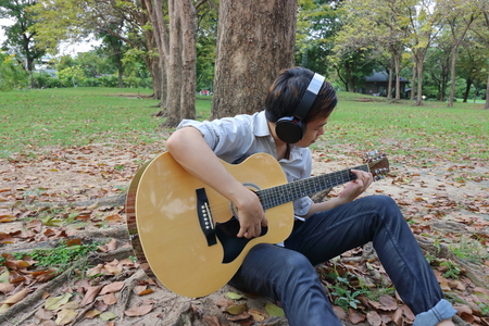 Portrait of happy young man playing acoustic guitar and leaning a tree at the outdoor park.の写真素材