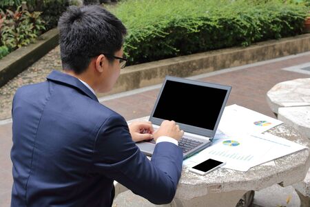 Rear view of handsome young business man is typing on the laptop against smartphone and documents on the top in public outdoors park.の写真素材
