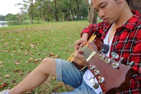 Close up young asian man in red shirt leaning against a tree and playing acoustic guitar in...