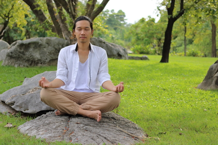 Young yogi man doing yoga meditation while sitting in lotus position on the rock in the park.の写真素材