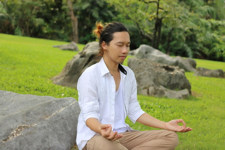 Portrait of young Asian man doing yoga meditation while sitting in lotus position on the rock in beautiful outdoor park.の写真素材