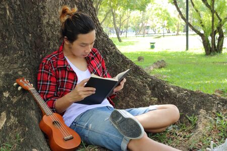 Young handsome man sitting on a green grass and reading a book in park.の写真素材