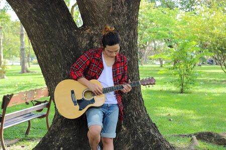 Portrait of young hipster guy standing against a tree and playing acoustic guitar in beautiful nature background.の写真素材