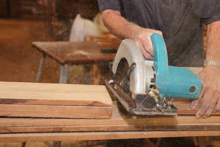 Selective focus on hands of senior carpenter cutting a piece of wood with electric circular saw in...