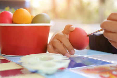 Close up hands of child coloring eggs with paintbrush for preparing Easter day on nature blurred background. Sunshine effect.の写真素材