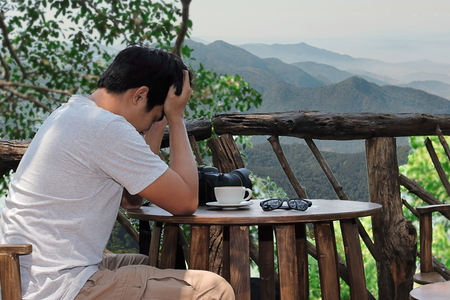 Stressed young Asian man depress with hands on forehead at coffee shop against nature mountain background.の写真素材