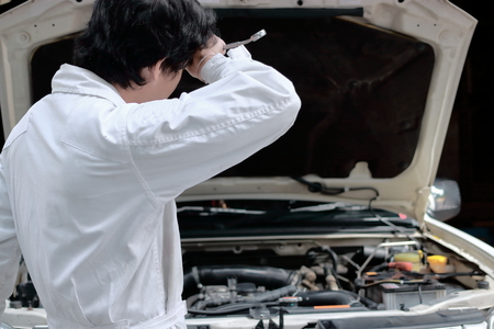 Portrait of tired young Asian mechanic man in uniform with wrench with car in open hood at the...