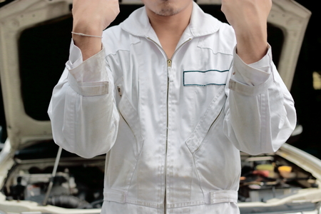Portrait of successful mechanic man in uniform raising hands up with car in open hood at the repair garage background.の写真素材
