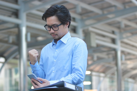 Successful young Asian business man looking mobile smart phone and feeling confident in outside office background.の写真素材