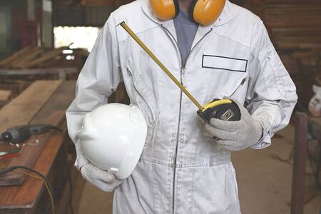 Portrait of young wood worker in white uniform holding safety helmet and measuring tape in carpentry workshop.の写真素材
