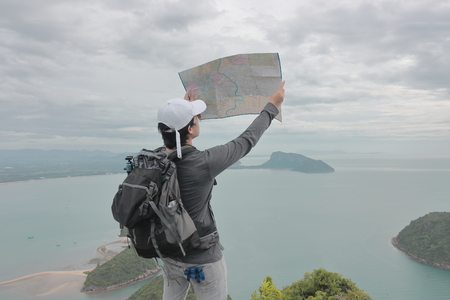 Young Asian hipster man with bag exploring map on viewpoint of mountain. Travel and adventure concept.の写真素材