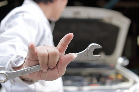 Back view of car mechanic man holding wrench and his hand with i love you sign with car in open hood at the repair garage background.の写真素材