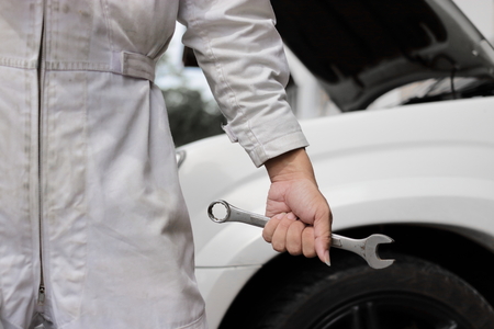 Hands of professional mechanic man in white uniform holding wrench with car in open hood at the repair garage background.の写真素材