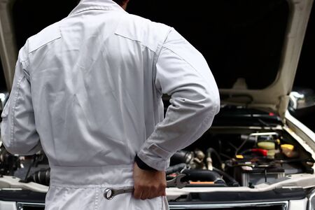 Back view of automotive mechanic in white uniform with wrench diagnosing engine under hood  at the repair garage. Car insurance concept.の写真素材