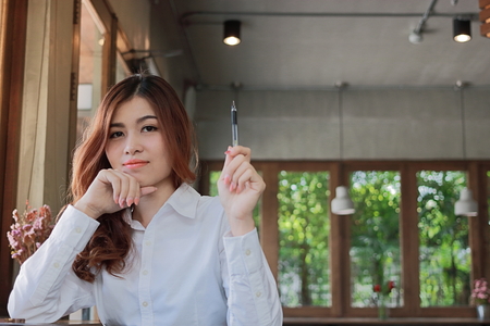 Portrait of attractive young Asian businesswoman holding a pen in her hands and looking at camera in coffee shop with copy space background.の写真素材