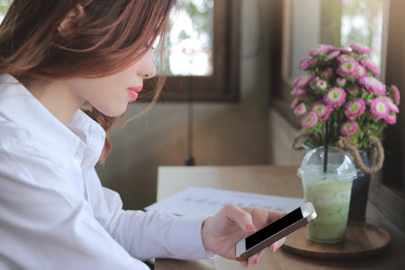Side view of attractive young Asian business woman looking mobile smart phone in her hands in coffee cafe.のeditorial素材