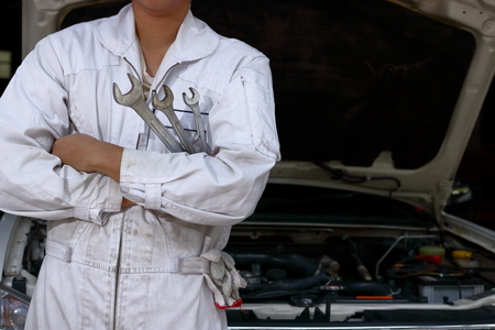 Portrait of professional young mechanic man in uniform holding wrench against car in open hood at the repair garage.の写真素材