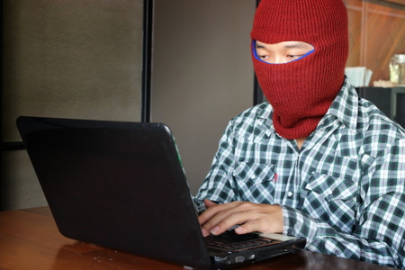 Masked hacker wearing a balaclava stealing data from laptop. Internet crime concept.