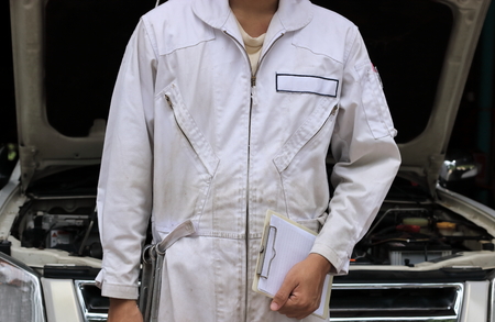 Professional young auto mechanic in uniform holding clipboard against car in open hood at the repair garage. Car insurance concept.の写真素材