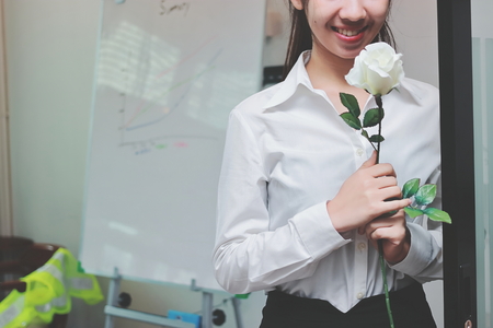 Vintage toned image of cheerful young Asian business woman holding a white roses in office on Valentine's day. Love and romance in workplace concept.の写真素材