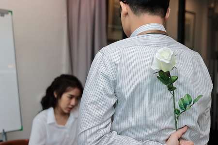 Young Asian business man holding a white rose behind his back for surprise girlfriend in valentines day. Love and romance in office concept.の写真素材