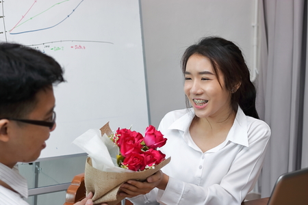 Cropped image of attractive young Asian woman accepting a bouquet of red roses from boyfriend in office on valentine's day. Love and romance in workplace concept.の写真素材