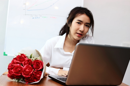 Vintage toned image of cheerful young Asian business woman with a bouquet of red roses on the desk in office. Valentine's day concept.の写真素材