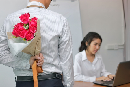 Young Asian business man holding a bouquet of red roses behind his back for girlfriend in valentines day. Love and romance in workplace concept.の写真素材