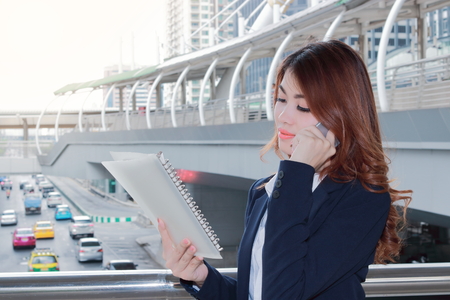 Portrait of pretty young Asian lady secretary talking on phone and looking paperwork in ring binder at urban city with sunshine effect background.の写真素材