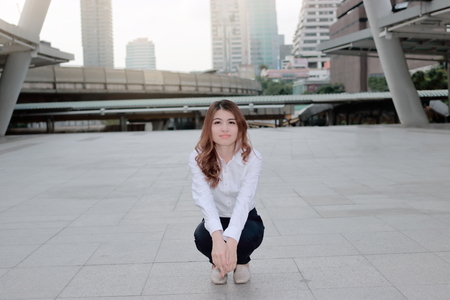Portrait of happy young Asian woman posing in a squatting position on sidewalk at urban city background.の写真素材