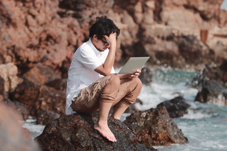 Stressed worried young Asian man with laptop sitting on the rock of sea shore suffering from severe depressionの写真素材