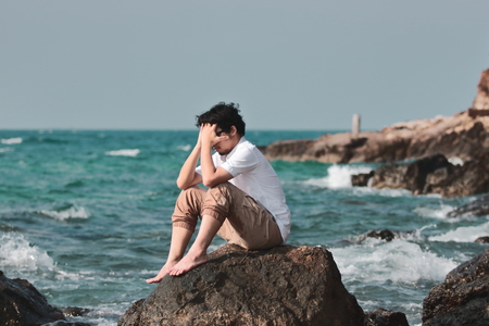 Portrait of lonely sad young Asian man covering face with hands and sitting on the rock of sea shoreの写真素材
