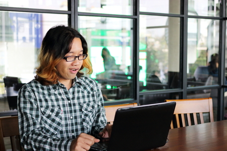 Cheerful young Asian hipster with casual clothes working with laptop for his job in workplace of...
