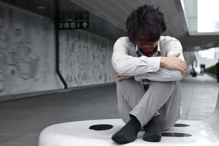 Frustrated exhausted young Asian business man sits and hugs his knees up to the chestの写真素材