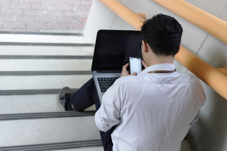 Back view of stressed young Asian business man sitting and using mobile smart phone and laptop at staircase of office.の写真素材
