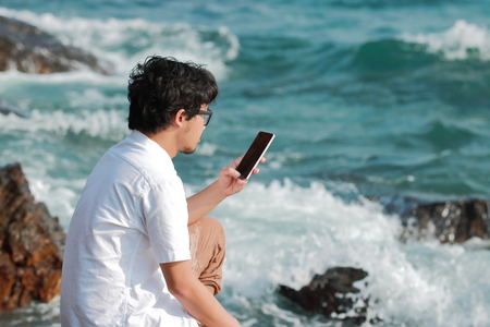 Young Asian man with mobile smart phone sitting on the rock of natural seashore. Summer lifestyle concept.の写真素材