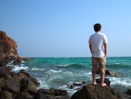 Back view of lonely Asian man standing on the stone of sea shore and looking at far away.の写真素材