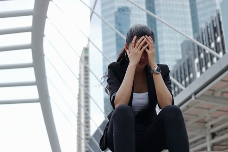 Depressed stressed young Asian business woman with hands on face sitting at outdoors.