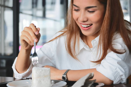 Cheerful Asian woman eating a peace of coconut cream cake in living roomの写真素材
