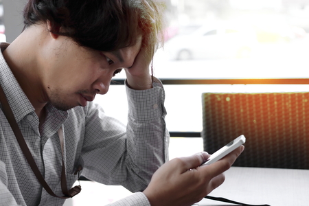 Frustrated stressed Asian business man with hands on forehead looking mobile smart phone in his hands at office.の写真素材