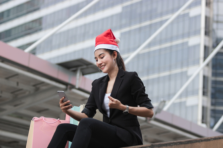 Cheerful young Asian woman with santa hat and colorful shopping bag using mobile smart phone for shopping online for christmas giftsの写真素材