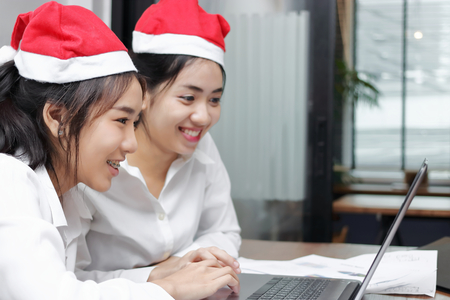Two cheerful young Asian business women with santa claus hat shopping online together in christmas holiday. Internet of thing concept.の写真素材