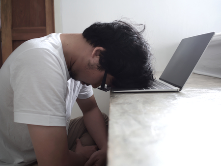 Tired upset young Asian man with head down on computer laptop.の写真素材