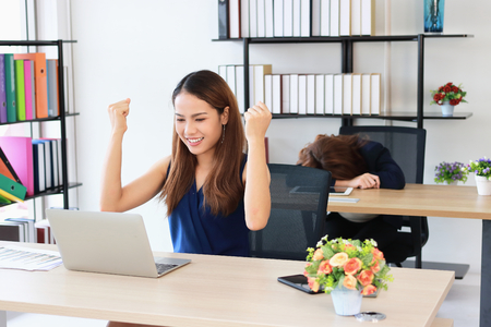 Successful Asian business woman raising hands with competitor colleague bend down head on the desk in office.の写真素材