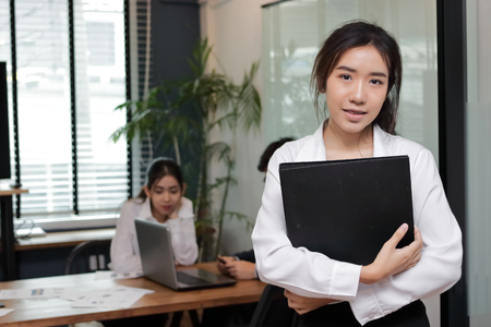 Leadership business woman concept. Cheerful young Asian businesswoman with ring binder standing against her colleague in office background.の写真素材
