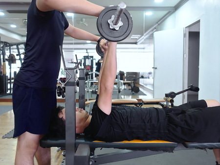 Healthy young Asian man lifting barbell with personal trainer at sport gym. Fitness and workout...
