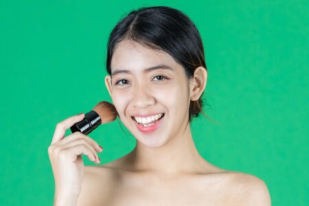 Beauty face of young Asian woman applying make up with brush over green isolated background. Healthy and cosmetics concept.の写真素材