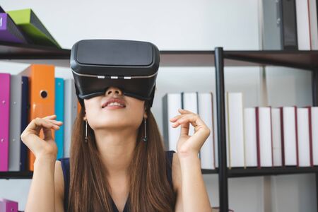 Relaxed young Asian woman wearing virtual VR reality glasses on workplace in office.