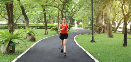 Panoramic view of relaxed smiling Asian fitness runner woman listening music and running workout in natural park.の写真素材
